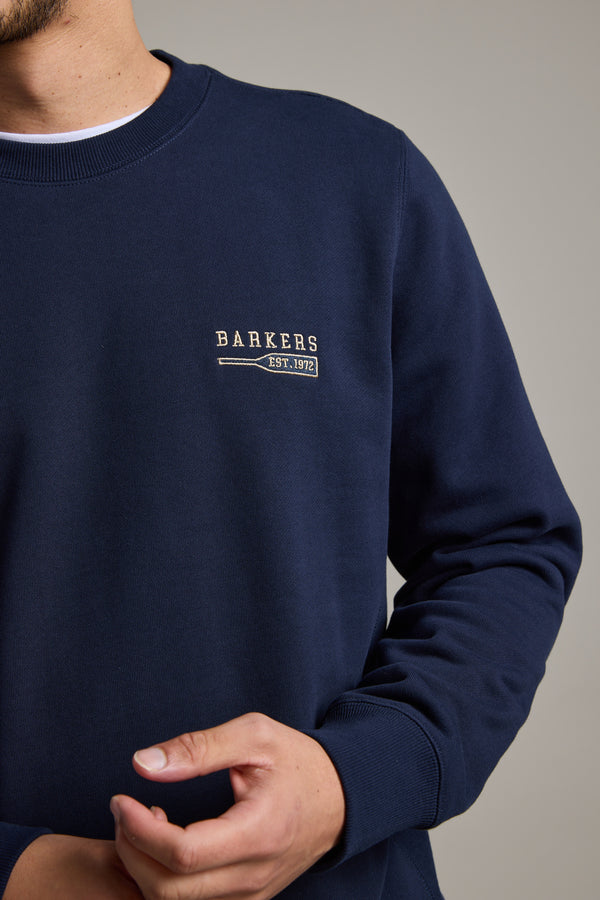 A person wears a navy Barkers Regatta Crew sweatshirt with "BARKERS EST. 1972" embroidered in white, made from responsibly sourced cotton, shown from shoulders to waist with one hand partially visible.