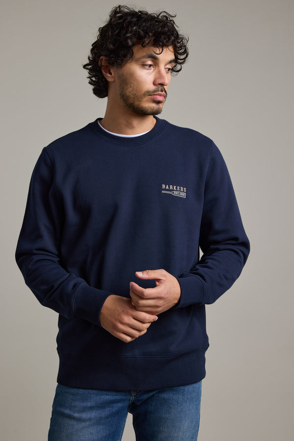 A man with curly hair and a short beard wears the Barkers Regatta Crew by Barkers—a navy crewneck sweatshirt made from responsibly sourced cotton with small white text on the left chest—paired with blue jeans, against a light gray background.