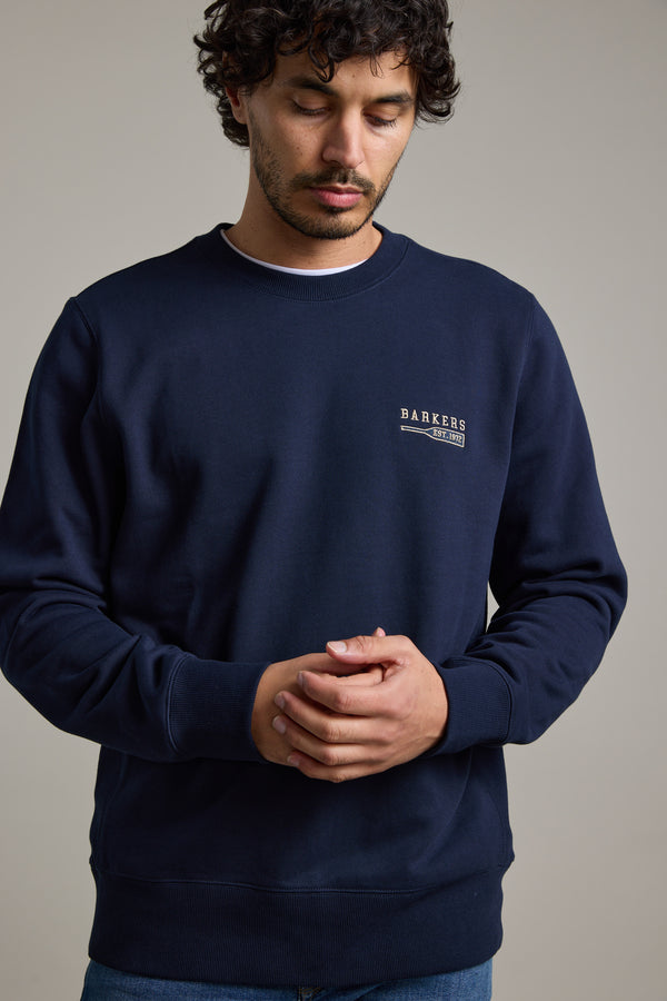 A man with curly hair wears the Barkers Regatta Crew sweatshirt by Barkers, made from responsibly sourced cotton, standing against a plain background and looking down with his hands clasped.