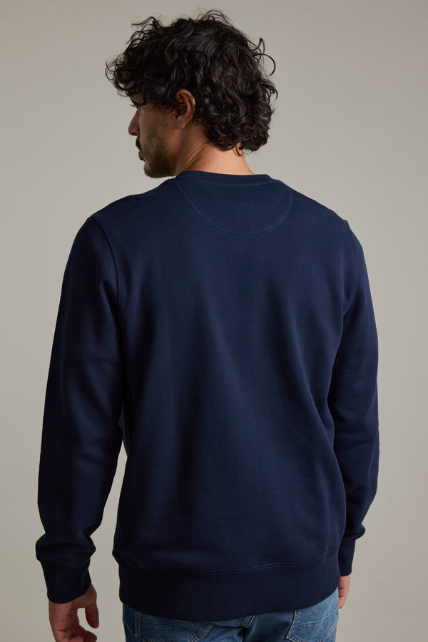 A person with curly dark hair faces away from the camera, wearing a Barkers Regatta Crew sweatshirt by Barkers and blue jeans, set against a plain light gray background.