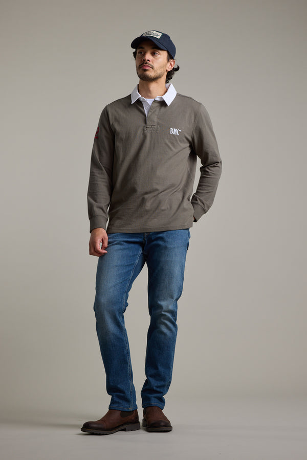A man wears a dark baseball cap, Barkers Stanford Long Sleeve Rugby in gray, blue jeans, and brown shoes. He stands against a plain light background with one hand in his pocket, looking slightly upward.