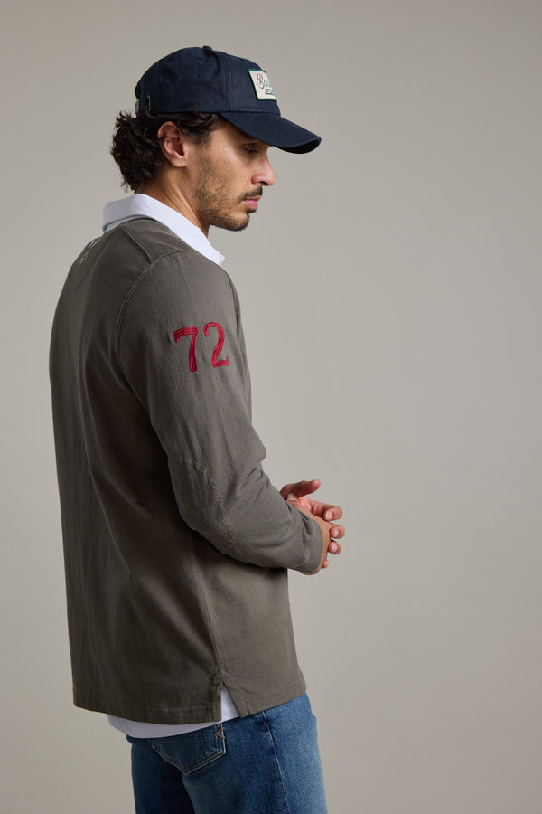A man in a Barkers Stanford Long Sleeve Rugby with an olive design and red 72 on the sleeve, navy cap, white undershirt, and blue jeans stands against a plain background, looking to the side with hands clasped.