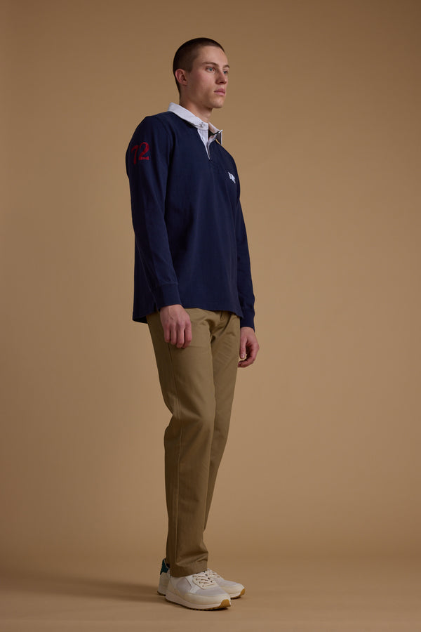 A man stands against a plain light brown background, wearing the Barkers Stanford Long Sleeve Rugby—navy with red 72 on the sleeve—paired with khaki pants and white sneakers. His hands rest at his sides as he looks forward.