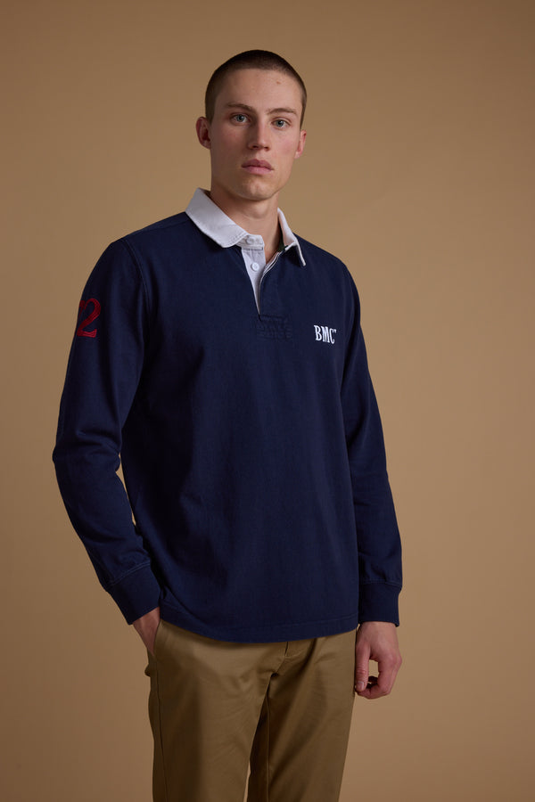 A young man with short hair wears the Barkers Stanford Long Sleeve Rugby in navy blue with a white collar and tan pants, standing before a plain brown background and looking directly at the camera.