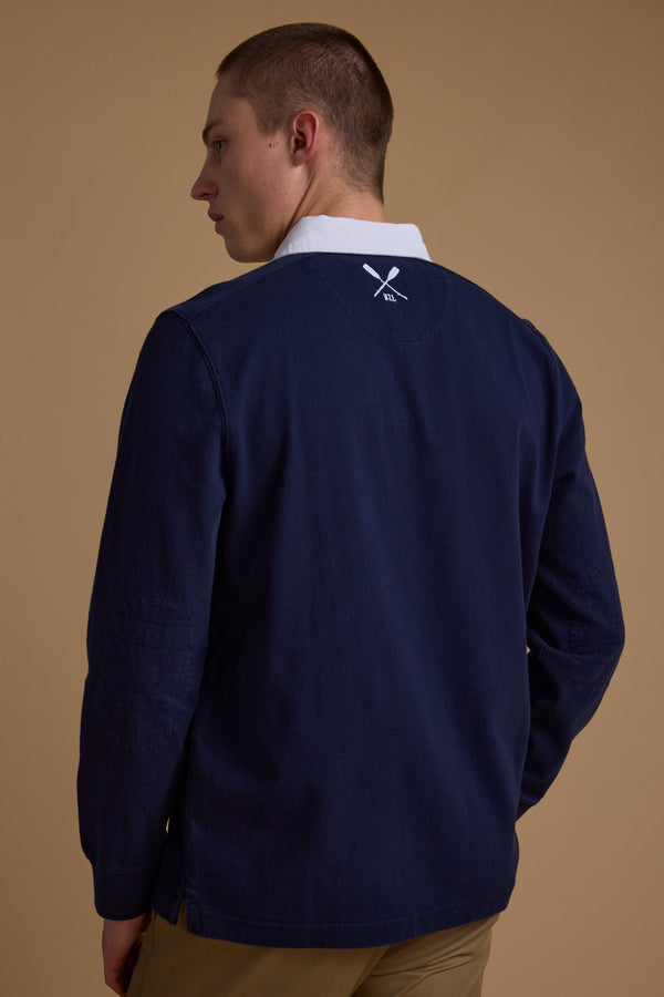 A man with short hair, wearing Barkers' Stanford Long Sleeve Rugby in navy with a white collar and khaki pants, stands facing away on a beige backdrop. A white crossed paddles logo appears on the back of his jersey.