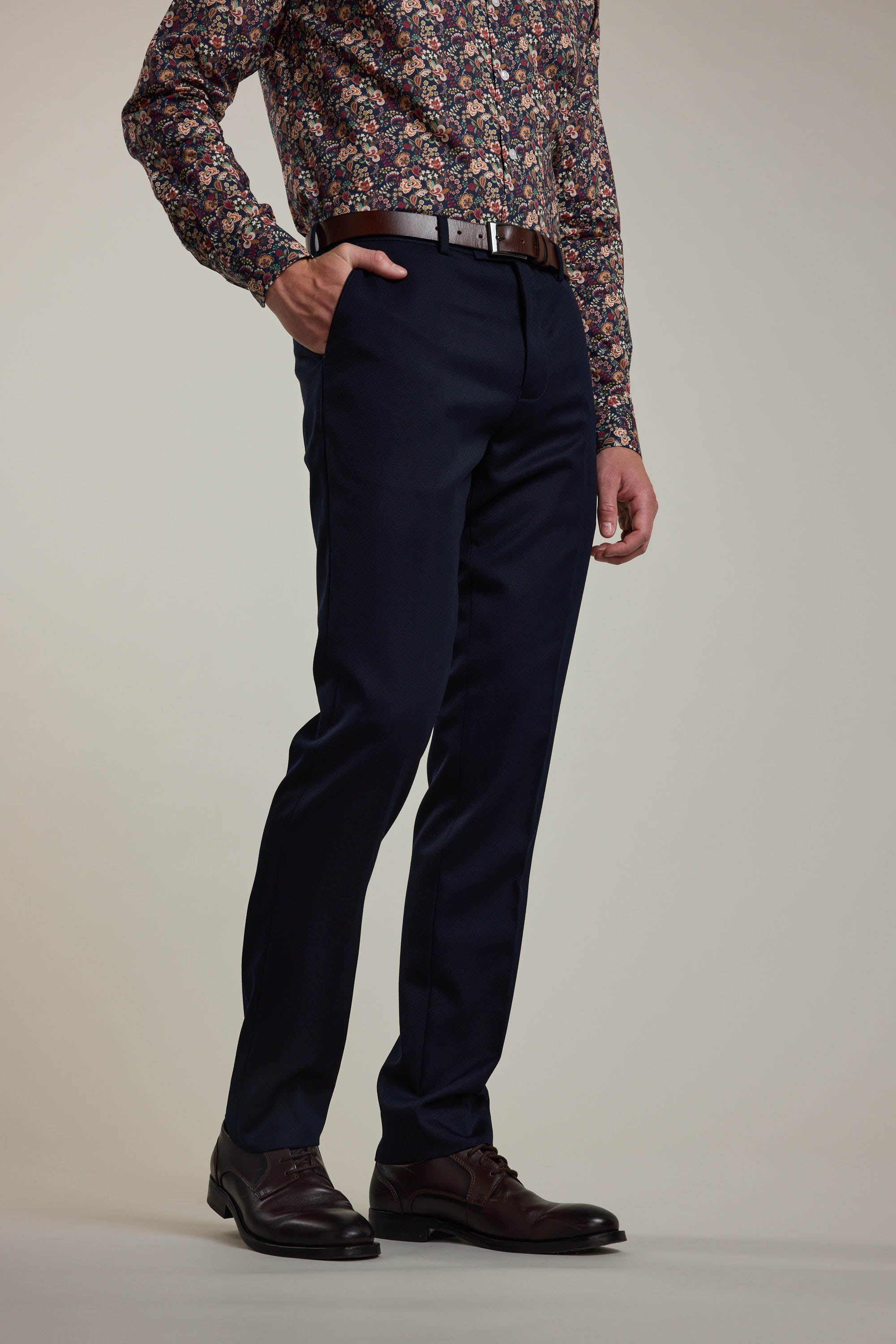 A person, with one hand in their pocket, wears Barkers Croydon Twill Suit Trouser in navy blue, a brown belt, brown shoes, and a long-sleeve shirt with a colorful floral pattern. The upper part of the head is not visible.