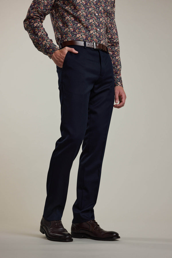 A person, with one hand in their pocket, wears Barkers Croydon Twill Suit Trouser in navy blue, a brown belt, brown shoes, and a long-sleeve shirt with a colorful floral pattern. The upper part of the head is not visible.