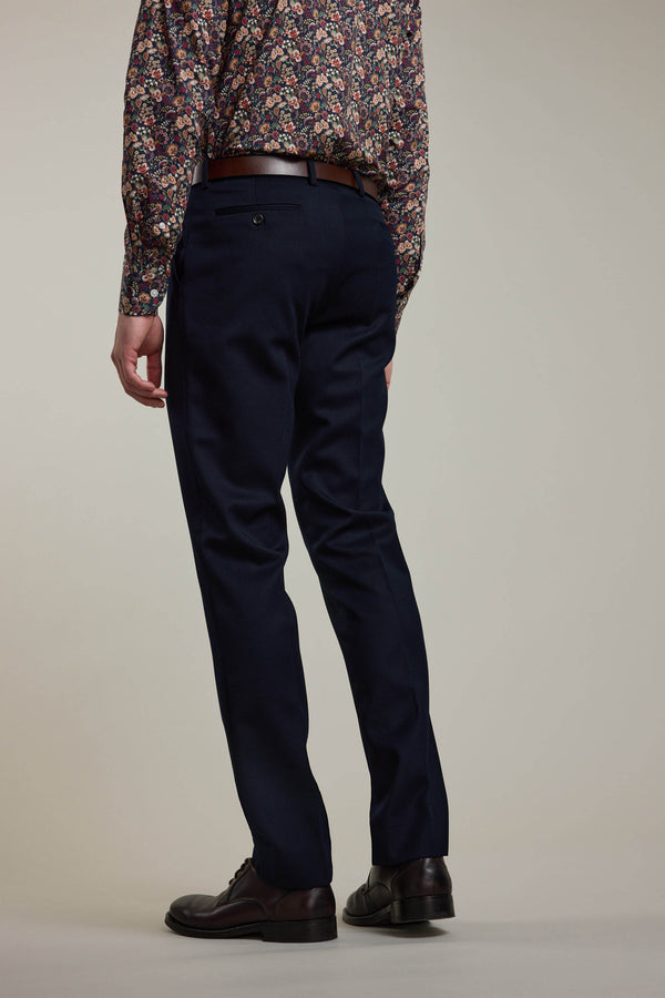 A person stands with their back to the camera, wearing Barkers Croydon Twill Suit Trouser in dark navy, paired with a brown belt, black shoes, and a long-sleeve floral shirt. The background is plain and neutral.