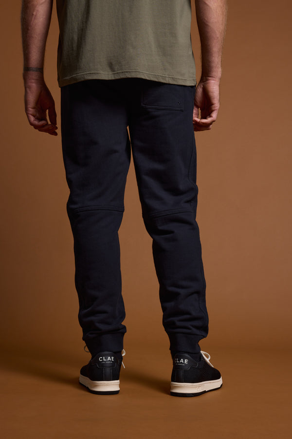 A person stands facing away against a brown background, wearing a green T-shirt, Barkers Heritage Track Pant by Barkers in dark color, and black sneakers with white CLAE branding on the soles.