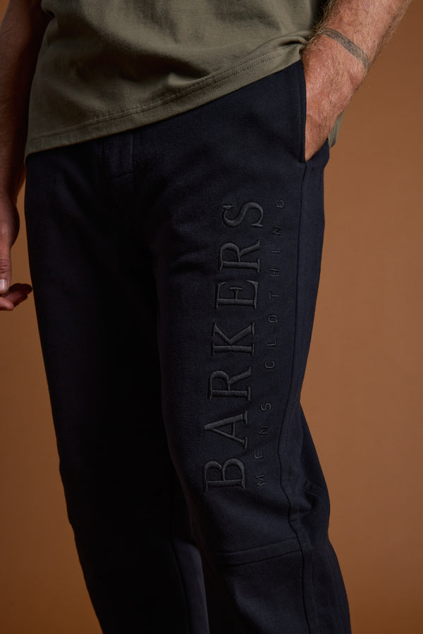 A person in Barkers Heritage Track Pant, featuring the Barkers logo on the side, with an olive-green shirt. Only their lower torso and one hand are visible against a brown background.