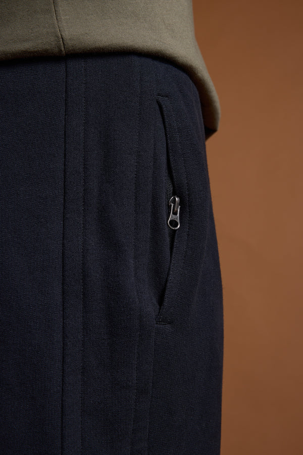 Close-up of a person wearing Barkers Heritage Track Pant by Barkers in dark navy with a zippered pocket, paired with an olive green top against a brown background.