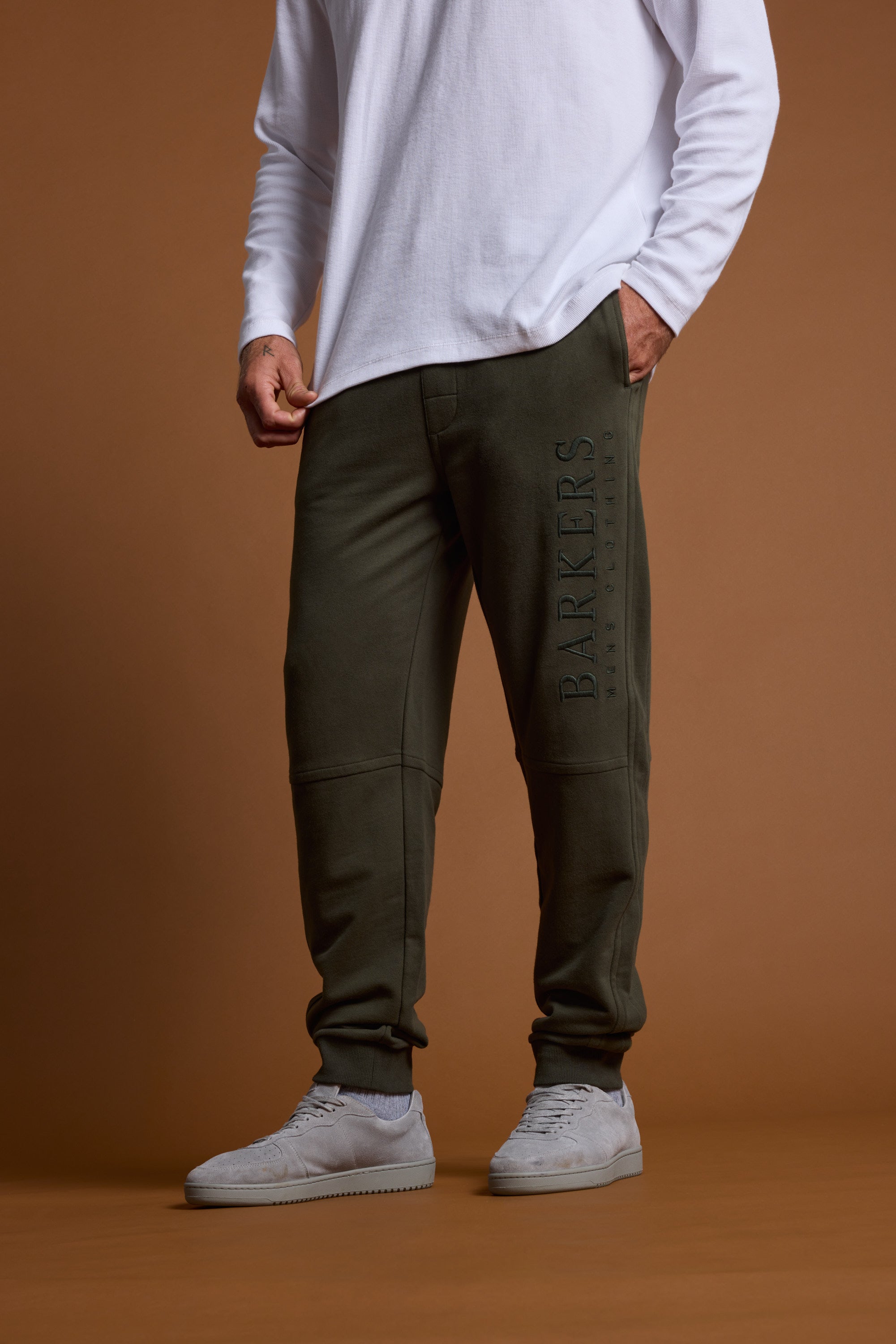 A person stands against a plain brown background in olive green Barkers Heritage Track Pants with "BARKERS" on the left leg, a long-sleeved white shirt, and light gray sneakers.