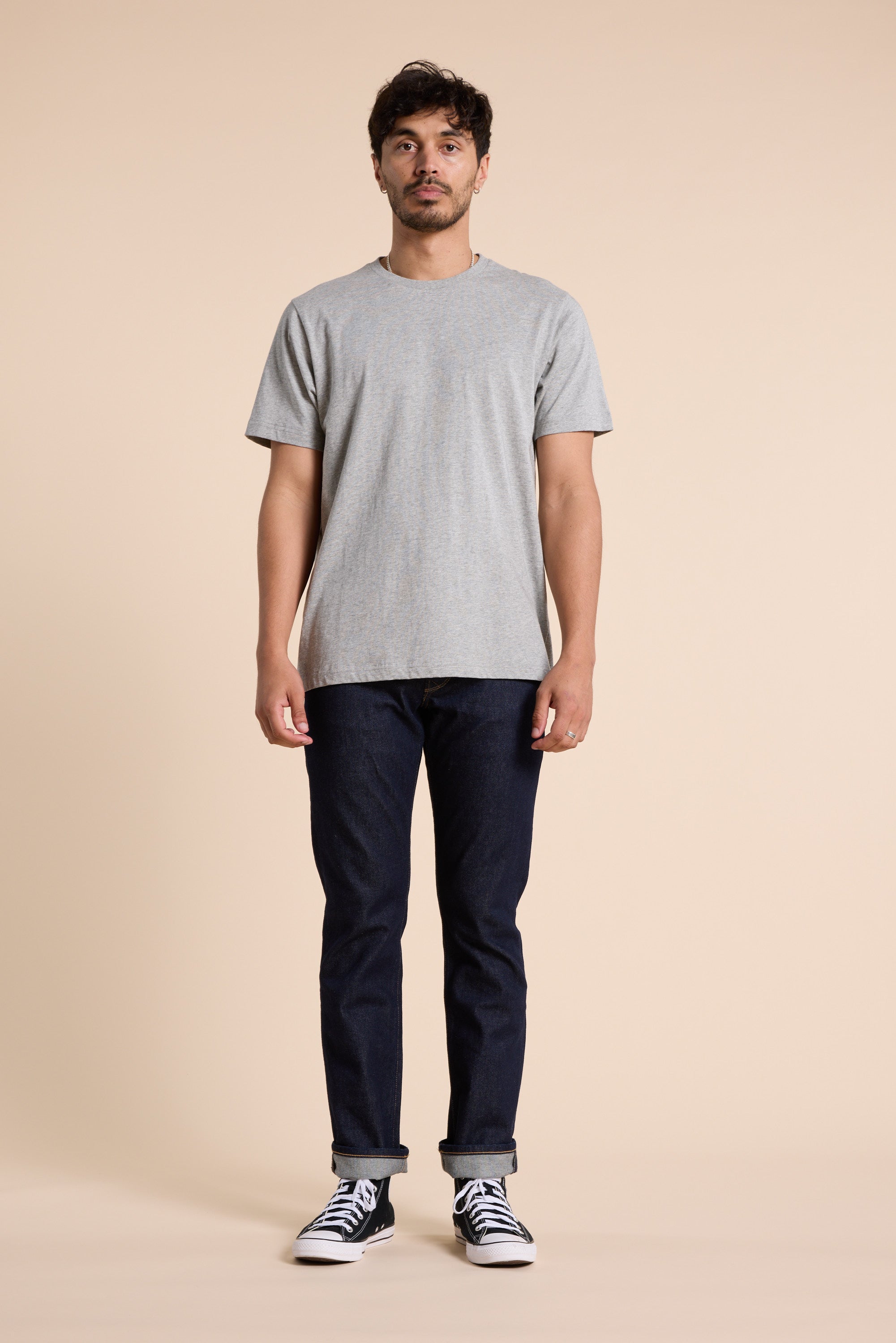A man stands facing forward against a beige background, wearing a Barkers Organic Crew Tee in light gray made from organic cotton, paired with dark blue jeans and black-and-white sneakers. His arms rest naturally by his sides.