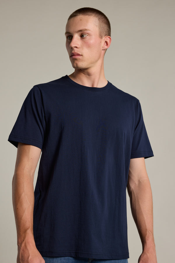 A young man with short hair wears a Barkers Organic Crew Tee in plain navy blue made from organic cotton, paired with blue jeans, standing against a neutral background and looking slightly to his left.