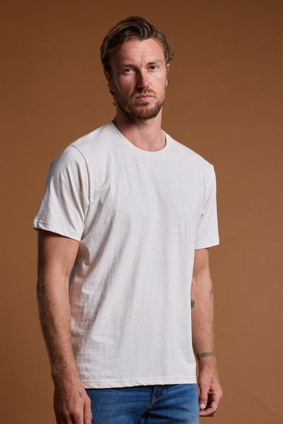A man with short, wavy hair and a beard stands against a plain brown background, wearing the Barkers Organic Crew Tee made from 100% organic cotton and blue jeans, looking directly at the camera with a neutral expression.