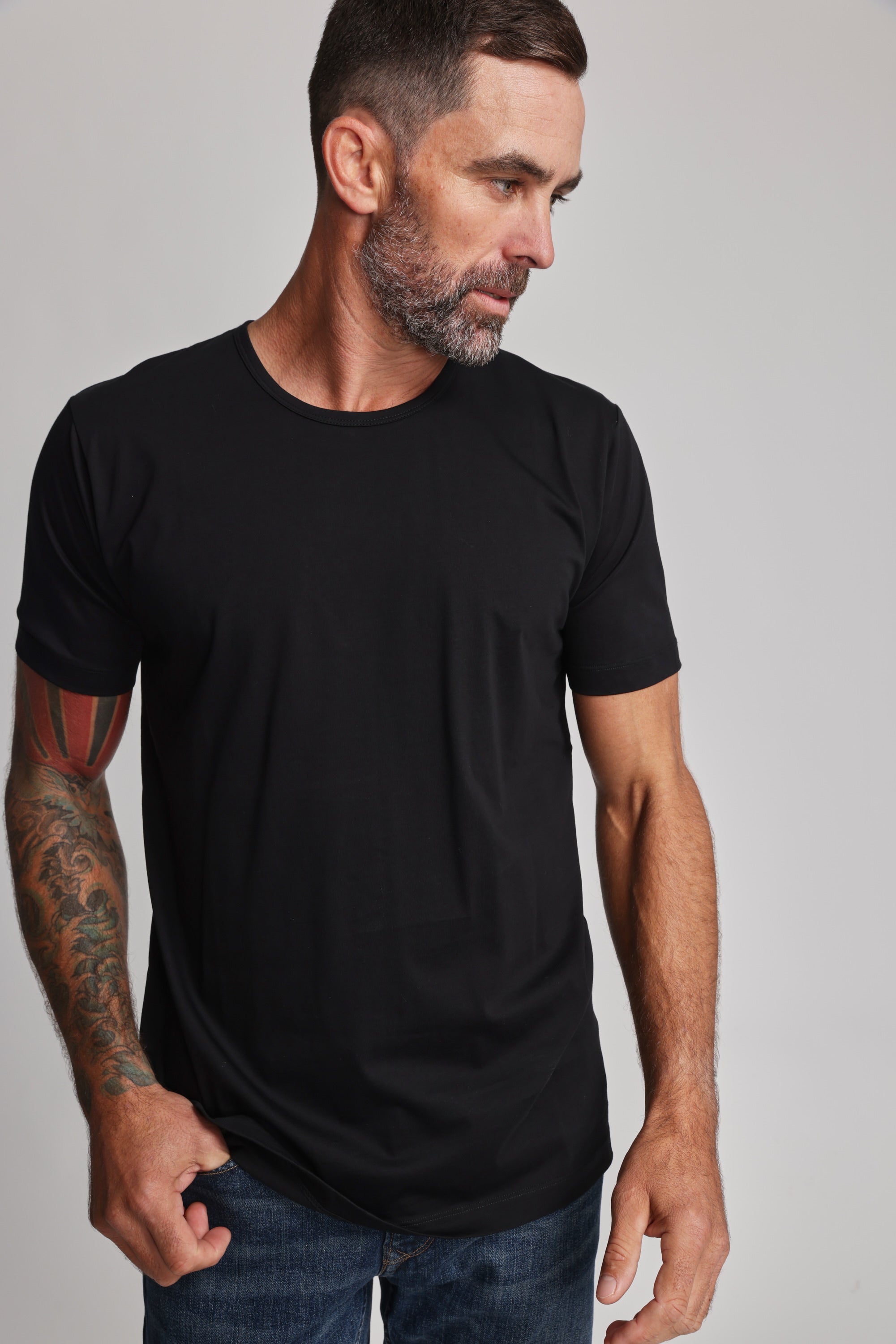 A man with short dark hair, a beard, and tattoos on his left arm wears the Barkers Linton Supima Tee in black with blue jeans. He looks down to his left against a plain light background.