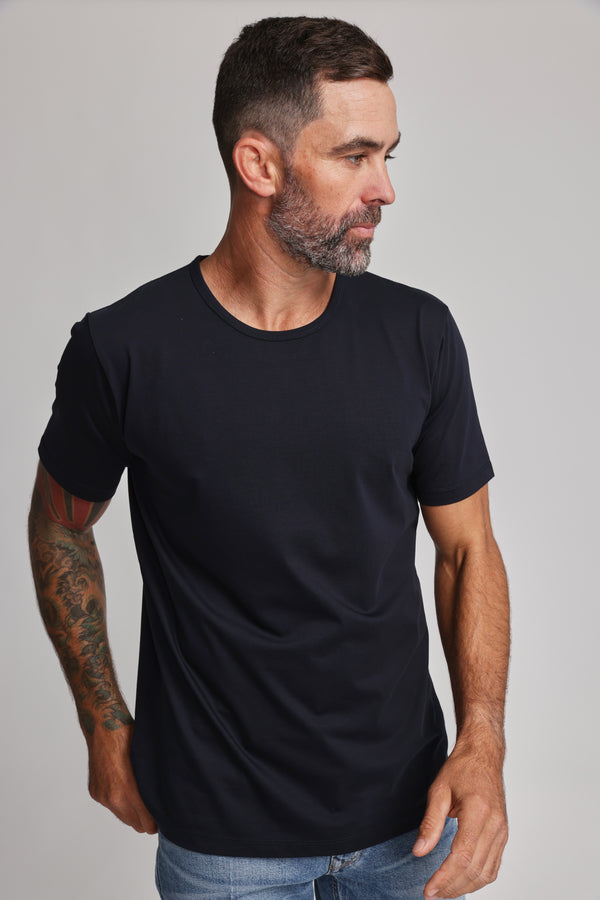 A man with short dark hair and a beard, wearing the Barkers Linton Supima Tee in black and light blue jeans, stands against a neutral background looking left. A colorful tattoo is visible on his arm—an ideal wardrobe staple.