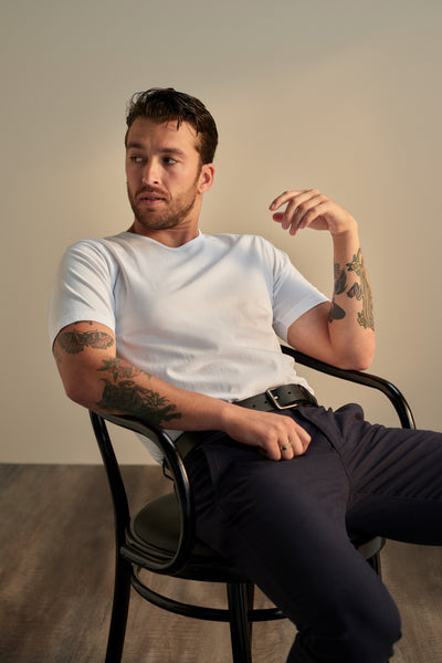 A tattooed man sits on a black chair in dark pants and the white Linton Supima Tee from Barkers. Relaxed, he looks to the side with one arm on the chair’s back and the other on his leg—a true wardrobe staple.