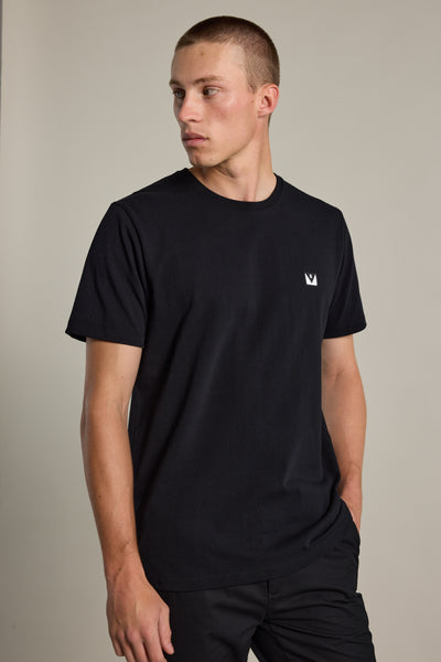 A person wearing the Barkers Newton Washed Tee, an oversized black box fit t-shirt with a small white crown logo on the chest, stands against a neutral background, looking to the side with one hand in their pocket.