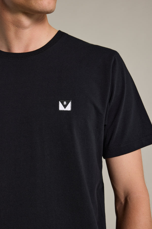 A person wearing the Newton Washed Tee by Barkers—an organic cotton black t-shirt with a small square "B" patch on the left chest. The person's face is partially out of frame.