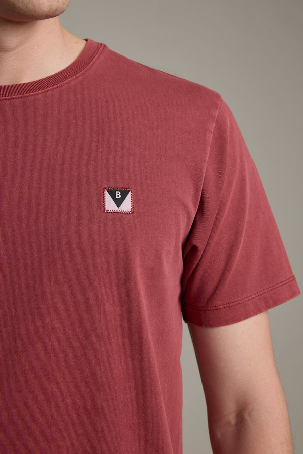 A person wears the Barkers Newton Washed Tee in faded red, featuring a small black and pink triangle "B" patch on the left chest, shown from shoulders to mid-torso against a plain background.