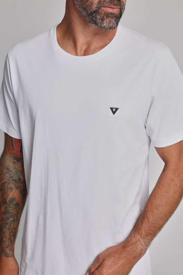 A man with a beard and tattooed arm wears the Barkers Newton Washed Tee, an organic cotton t-shirt in plain white with a small black and white geometric logo on the left chest. His face is partly out of frame, hand resting on his hip.