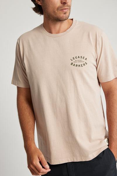 A man wears a light beige Legasea Marlin Print Tee by Legasea, made from organic cotton with short sleeves, a relaxed fit, and "LEGASEA BARKERS" printed on the left chest, standing against a neutral background.