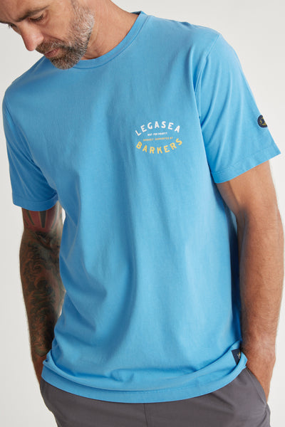 A bearded man wears the Legasea Fish For The People Tee by Legasea, a light blue organic cotton t-shirt with "LEGASEA BARKERS" on the chest, short sleeves, and a small black patch on the left sleeve. He looks down with one hand in his pocket.