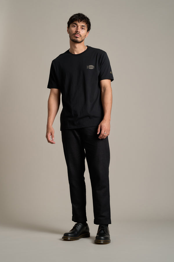 A man with short dark hair and slight facial hair stands against a beige background wearing the LegaSea Crayfish Print Tee by Legasea, paired with black pants and shoes. He has a relaxed posture with one hand by his side.