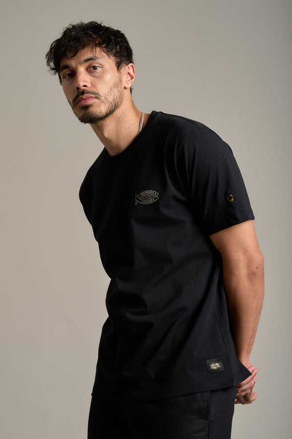 A man with short, curly dark hair and facial hair wears a Legasea Crayfish Print Tee featuring a small fish logo on the chest, standing against a beige background and showing support for New Zealand marine conservation.