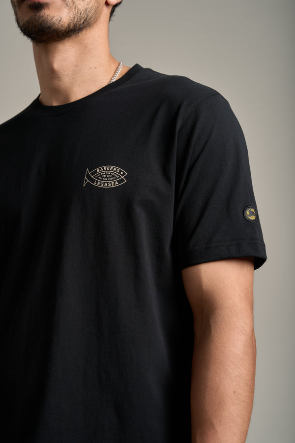 A person stands against a neutral background, wearing the Legasea Crayfish Print Tee—an organic cotton t-shirt with a small fish logo and text on the chest plus a circular patch on the sleeve. The look supports New Zealand marine conservation. Face partially out of frame.