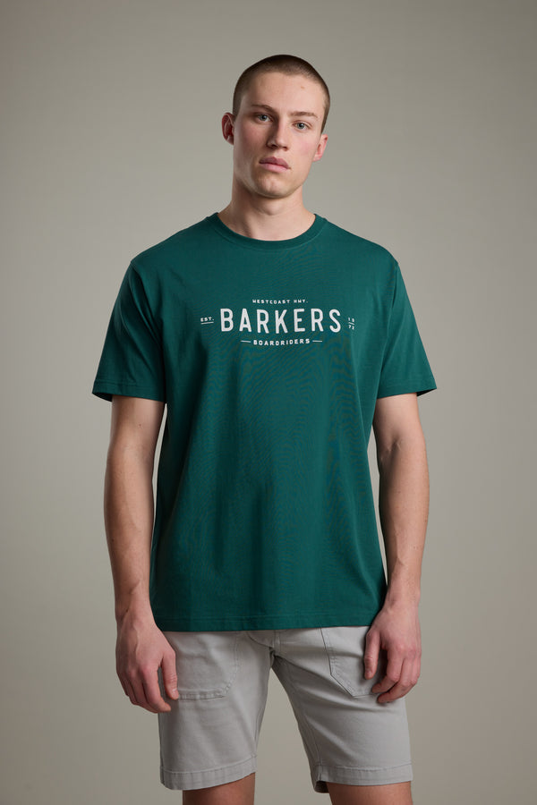 A young man stands against a light gray background, wearing the Barkers Boardriders Logo Tee in green and light gray shorts for an essential everyday look.