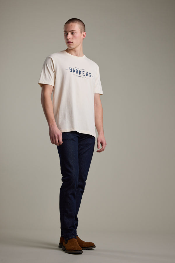 A man stands in a studio wearing a Barkers Boardriders Logo Tee, dark blue jeans, and brown suede boots, looking off to the side with a neutral expression against a plain background.