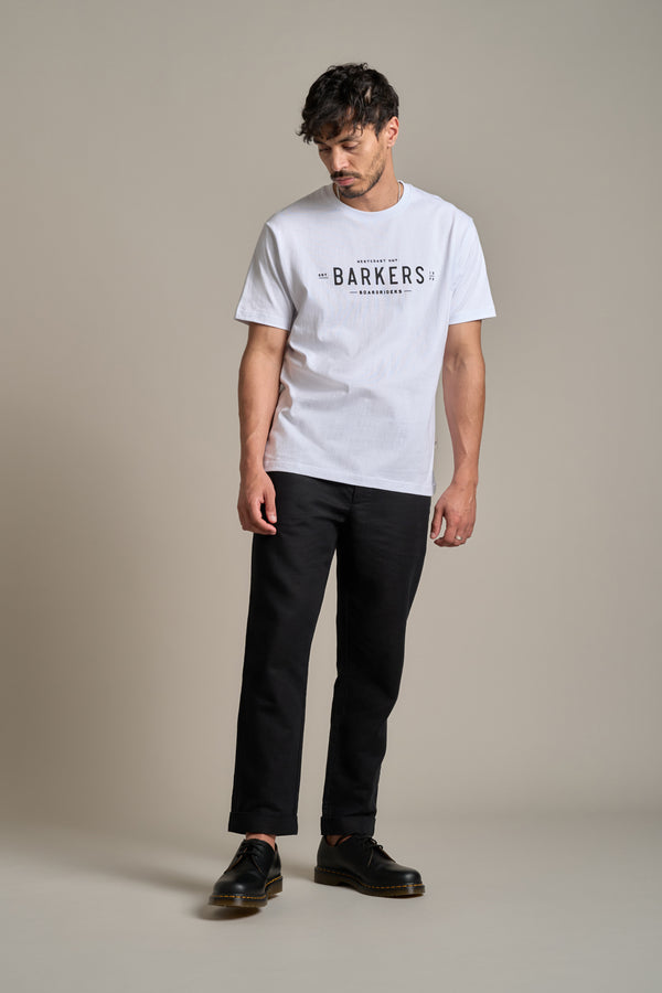 A man stands against a plain background wearing the Barkers Boardriders Logo Tee in white, paired with black pants and shoes. With his head slightly down and arms relaxed, he showcases a laid-back, essential everyday look.