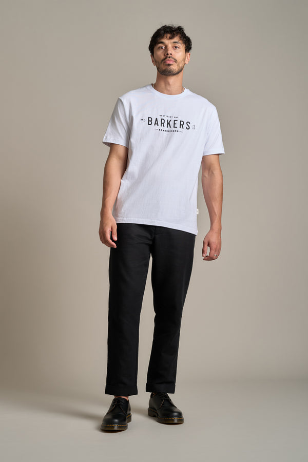 A man with short dark hair and a slight beard stands against a neutral background, wearing the Barkers Boardriders Logo Tee—an everyday essential organic cotton t-shirt—paired with black pants and shoes. His arms are relaxed at his sides.