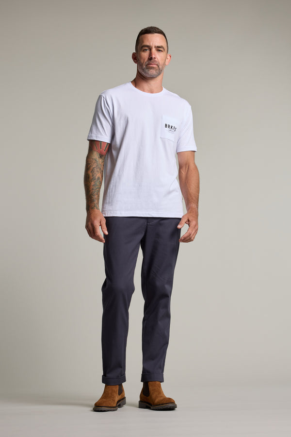 A man with short hair and right arm tattoos stands against a plain background, wearing a Barkers Basecamp Logo Tee by Barkers, dark gray pants, and brown suede boots—a staple for any Kiwi wardrobe.