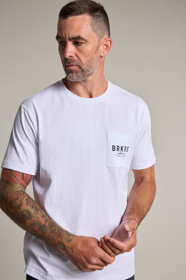 A man with short hair and a beard wears a Barkers Basecamp Logo Tee with a front pocket labeled BRKRS. He has a tattoo sleeve on his left arm, looking slightly to his right against a plain background. Brand: Barkers.