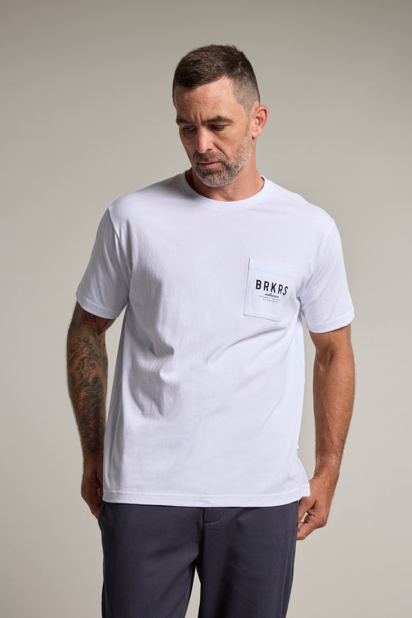 A man with short hair and a beard wears the Barkers Basecamp Logo Tee by Barkers, made from organic cotton with BRKPS on the chest pocket. He has a tattoo on his left arm and stands against a plain, light background.