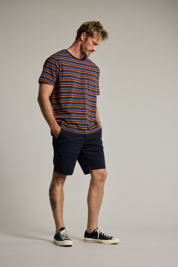 A man stands against a plain background in the Barkers Oxton Stripe Tee, crafted from organic cotton. He pairs it with navy shorts and black sneakers, standing relaxed with hands in pockets and a tattoo visible on his left calf.