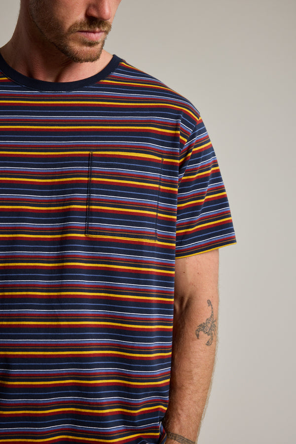 A person wears a Barkers Oxton Stripe Tee—men’s regular fit, organic cotton, multicolored stripes, chest pocket. Their lower face shows and a scorpion forearm tattoo is visible against a neutral background.