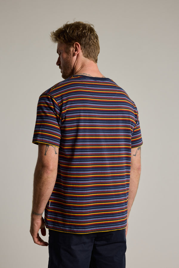 A man with light brown hair and tattooed arms stands with his back to the camera, wearing the Barkers Oxton Stripe Tee—a men's regular fit tee made from organic cotton with multicolored stripes—against a plain light background.