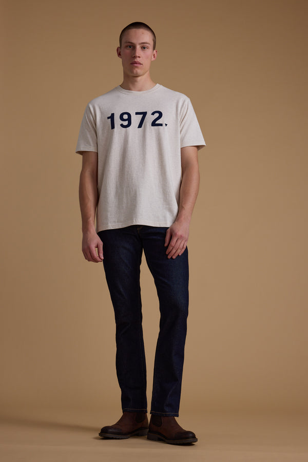 A man faces forward against a beige background, wearing the Barkers 1972 Print Tee made from organic cotton, paired with dark jeans and brown shoes.