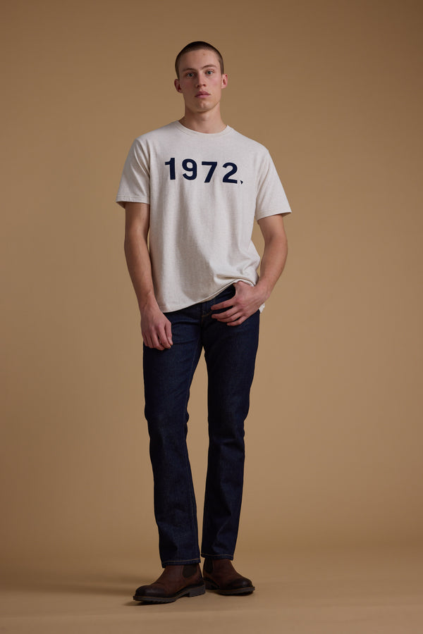 A person stands against a beige background, wearing a light gray Barkers 1972 Print Tee made from organic cotton, paired with dark jeans and brown shoes—one hand in their pocket for a classic menswear style.
