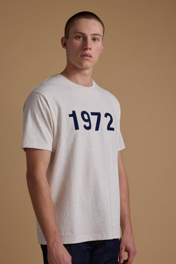 A young man with short hair stands against a beige background, wearing the Barkers 1972 Print Tee made from soft organic cotton, effortlessly showcasing modern menswear style.