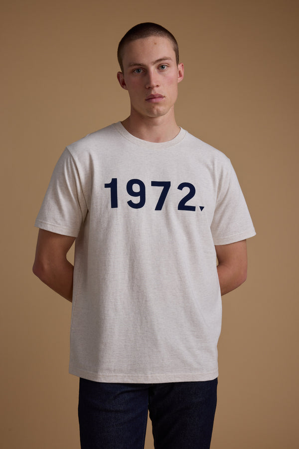 A young man stands against a beige background wearing the Barkers 1972 Print Tee—a light gray organic cotton shirt with bold navy numbers on the front. His hands are behind his back, and he has a neutral expression.