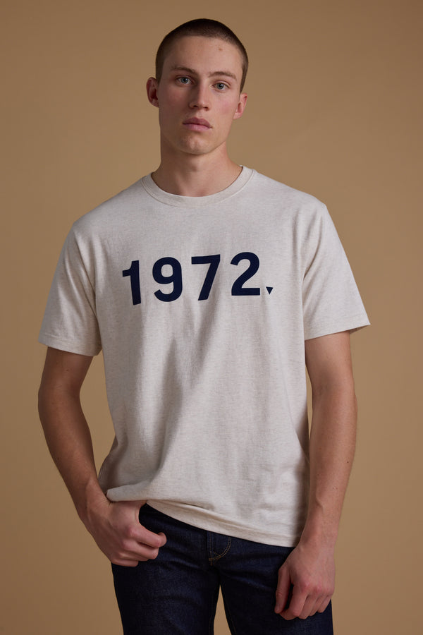 A young man with short hair wears the Barkers 1972 Print Tee, made from organic cotton and featuring bold navy numbers on the front. He stands against a taupe background, looking straight at the camera with a neutral expression.