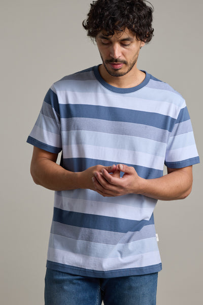 A man with curly hair and a short beard wears the Barkers Fitzroy Stripe Tee in blue and white stripes with jeans, looking down and touching his fingers against a plain light background.