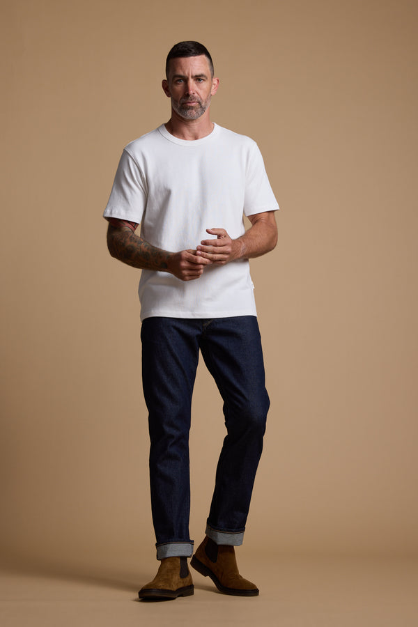 A man with short dark hair and a beard stands against a beige background, wearing the Barkers Langwood Tee, dark cuffed jeans, and brown suede boots. Tattoos are visible on his left forearm as he looks straight ahead.