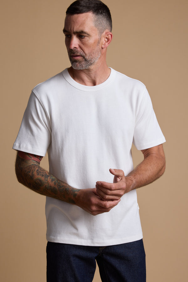 A man with short dark hair, a beard, and visible arm tattoos wears the Barkers Langwood Tee in a relaxed fit with dark jeans, standing against a beige background. He looks to his left while clasping his hands.