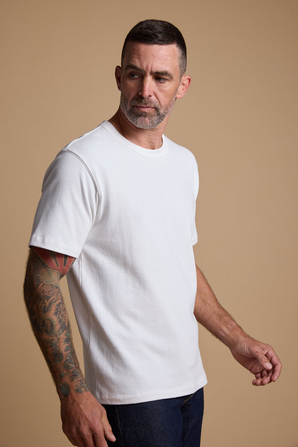 A man with short dark hair, a beard, and tattoos on his left arm stands against a beige background wearing the Barkers Langwood Tee in plain white and dark jeans. He is looking to his left.